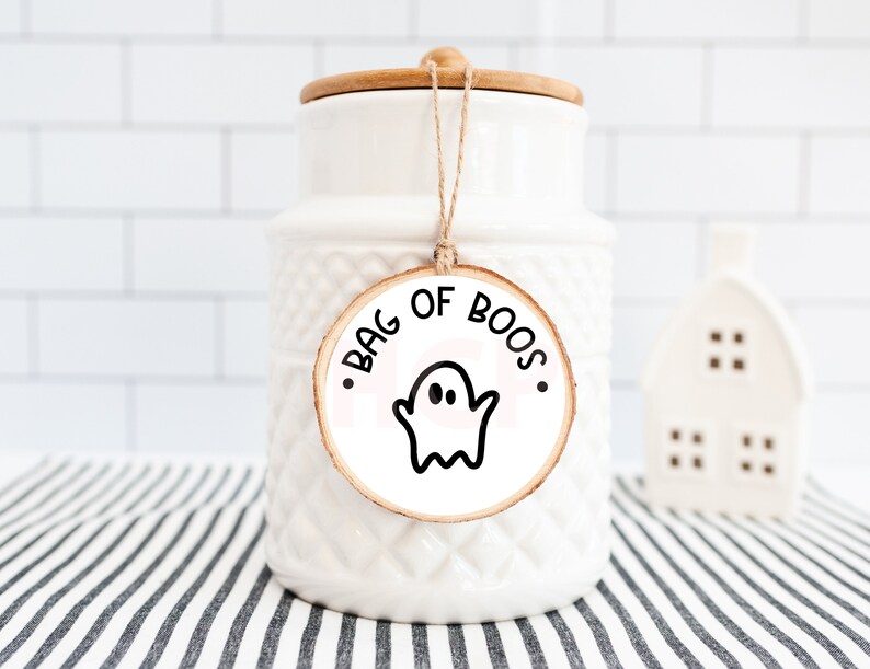 Pu&ograve; includere: Un barattolo di ceramica bianco con coperchio in legno e un'etichetta decorativa con la scritta "BAG OF BOOS" e un'illustrazione di un fantasma. Il barattolo &egrave; su una superficie a righe, con una piccola statuina bianca a forma di casa sullo sfondo.