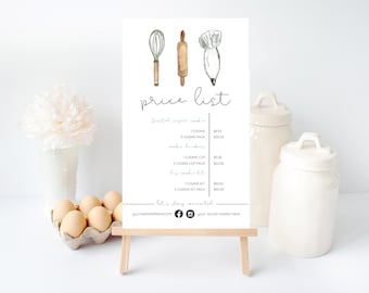 May include: A white sign with a price list for sugar cookies. The sign has a watercolor illustration of a whisk, rolling pin, and piping bag. The price list includes prices for individual cookies, cookie packs, cookie cups, and cookie kits.