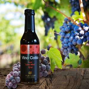 May include: A glass bottle of Vino Cotto Sweet Cooked Wine Syrup, with a red and black label, sitting on a wooden surface with grapes in the background.