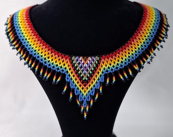 May include: A vibrant, beaded necklace displayed on a black mannequin. The necklace features a rainbow of colors, including red, orange, yellow, green, blue, and purple. The design includes a V-shaped pendant with dangling beads.