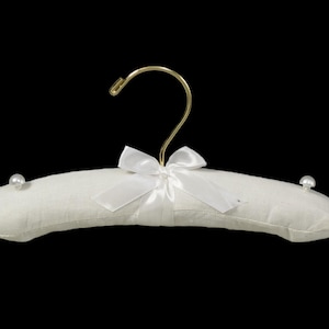 May include: White padded hanger with a gold hook and a white satin bow. The hanger has two pearl-like embellishments on the ends.