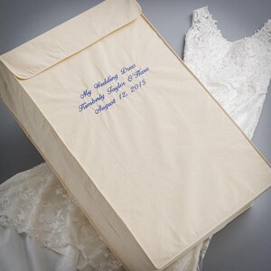 Op de afbeelding: Een crèmekleurige opbergdoos voor een trouwjurk met een klepsluiting. De doos heeft blauwe geborduurde tekst: "My Wedding Dress Kimberly Taylor O'Hara August 12, 2015." Een witte kanten trouwjurk is gedeeltelijk zichtbaar.