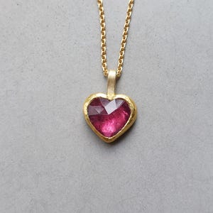 Può includere: Una collana in oro con un ciondolo a forma di cuore che presenta una gemma sfaccettata color rosa intenso, incastonata in una cornice d'oro testurizzata. Il ciondolo pende da una delicata catenina d'oro con maglie piccole e uniformi.