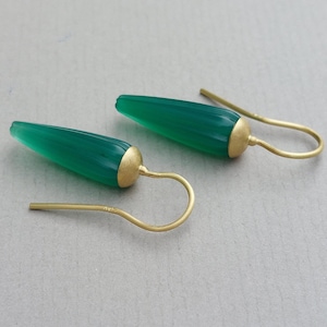 Op de afbeelding: Een paar oorbellen met langwerpige, taps toelopende groene stenen met een gladde, gepolijste afwerking. Elke steen is voorzien van een matte, goudkleurige metalen kap die verbonden is met een gebogen, goudkleurige oorhaak voor ophanging.