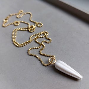 Op de afbeelding: Een goudkleurige kogelketting met een witte, langwerpige hanger. De hanger heeft een geribbeld ontwerp en is met een kleine gouden ring aan de ketting bevestigd. De ketting wordt op een grijze ondergrond getoond.