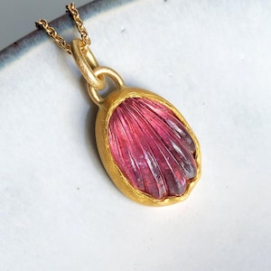 Può includere: Una collana con pendente caratterizzata da una gemma intagliata di colore rosa intenso, di forma ovale, incastonata in una montatura in metallo dorato opaco e testurizzato. Il pendente pende da una sottile catenina dorata con motivo a coste.