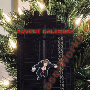 Puede incluir: Un calendario de adviento negro con una cuenta regresiva del 25 al 1. Un personaje de dibujos animados cuelga de la parte superior del calendario. El calendario está hecho de madera y tiene un acabado negro. El texto "ADVENT CALENDAR" está impreso en la parte superior del calendario.