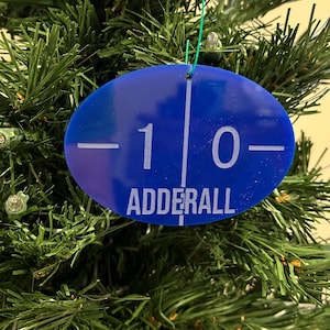 May include: A blue oval Christmas ornament with the word "ADDERALL" in white text. The ornament also has the numbers "-10-" in white. The ornament is hanging on a green Christmas tree.