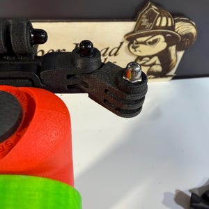 May include: A black camera mount with a red and green base. The mount has multiple connection points and a silver bolt. A wooden sign with a squirrel wearing a firefighter's helmet is in the background.