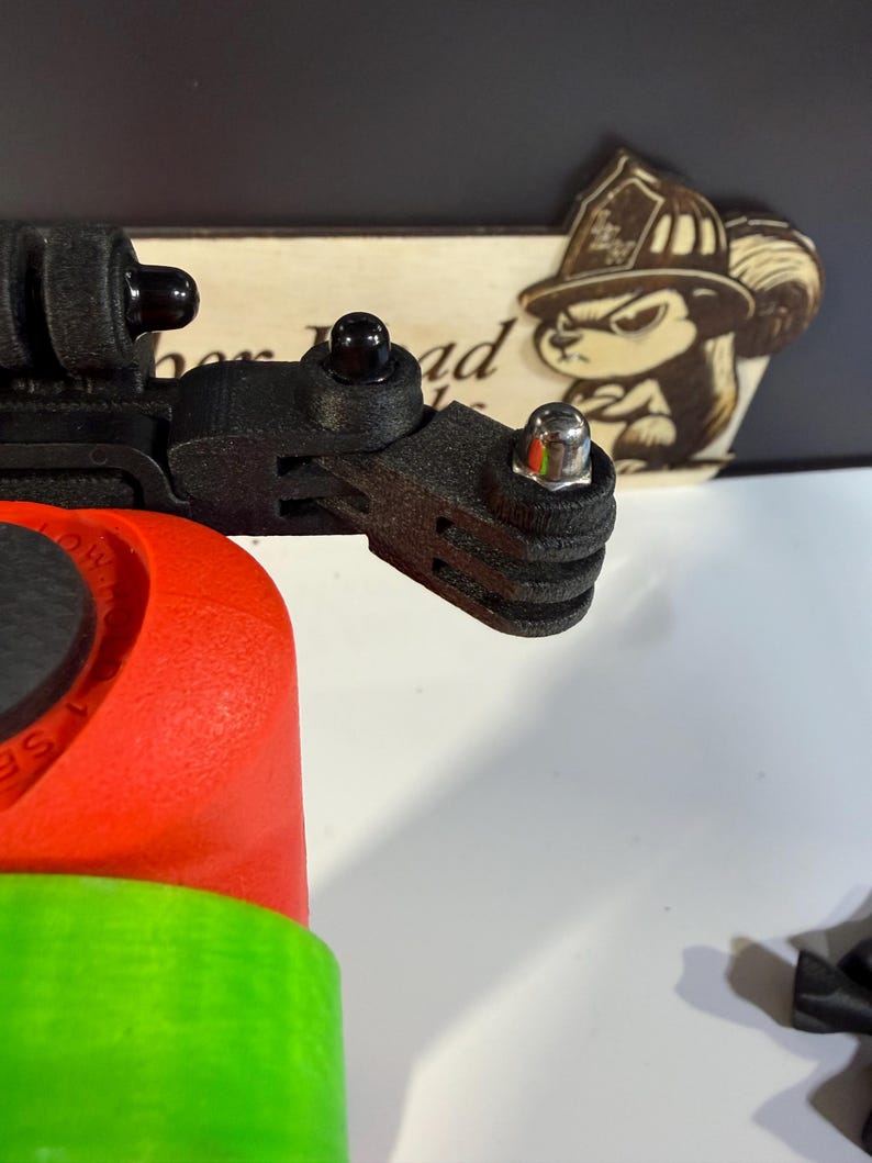 May include: A black camera mount with a red and green base. The mount has multiple connection points and a silver bolt. A wooden sign with a squirrel wearing a firefighter's helmet is in the background.