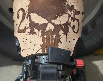 May include: A brown leather mask cover with a white skull graphic and the numbers "25" on either side. The cover is designed to fit a Scott AV3000 mask. The text "Custom Leather Scott AV3000 Mask Cover" is visible at the top of the image. The text "www.leatherheadlaserworks.com" is visible at the bottom of the image.