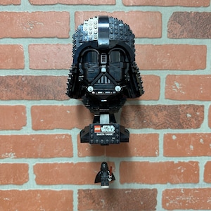 Op de afbeelding: Een zwarte LEGO Star Wars Darth Vader buste met een klein Darth Vader minifiguur dat eraan hangt. De buste is gemaakt van LEGO stenen en heeft een zwarte helm met een rood vizier. Het minifiguur houdt een rood lichtzwaard vast.