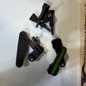 May include: A selection of black camera mounting accessories, including screws and brackets. One bracket has the text "LEATHERHEADLASERWORKS" in green. The items are arranged on a white surface.