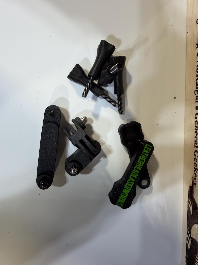 May include: A selection of black camera mounting accessories, including screws and brackets. One bracket has the text "LEATHERHEADLASERWORKS" in green. The items are arranged on a white surface.