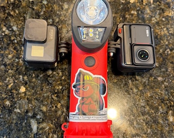 May include: A red flashlight with a bright LED light and a sticker of a firefighter squirrel. Two black action cameras are attached on either side. The flashlight has the text "LeatherHeadLaserWorks.com".