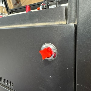 May include: A close-up of a red safety switch on a black metal panel. The switch is round with a star-shaped button.
