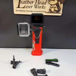 May include: A red action camera mount with a clear protective case and a black camera attached. The mount has a firefighter-themed design. Additional black mounting accessories are included. A wooden sign with the text "Leather Head Laser Works" is in the background.
