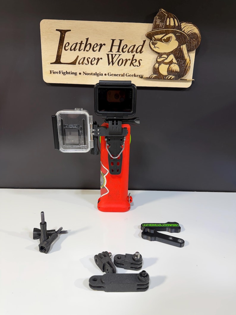 May include: A red action camera mount with a clear protective case and a black camera attached. The mount has a firefighter-themed design. Additional black mounting accessories are included. A wooden sign with the text "Leather Head Laser Works" is in the background.