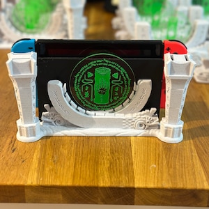 May include: A white and black gaming console stand with a green circular design. The stand has a unique architectural design with pillars and a curved base. The console is black with blue and red controllers.