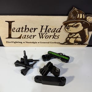 May include: A wooden sign with the text "Leather Head Laser Works" and a cartoon squirrel wearing a firefighter's helmet. Several black plastic mounting accessories are in the foreground, including brackets and screws. The sign also includes the words "FireFighting, Nostalgia, General Geekery."