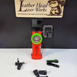 May include: A red flashlight with a green bezel and a GoPro camera attached. The flashlight has the word "SURVIVOR" printed on it. A wooden sign with the text "Leather Head Laser Works" and a cartoon squirrel is above the flashlight. Several black mounting accessories are in front.