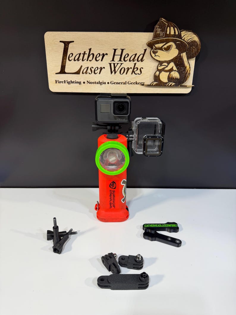 May include: A red flashlight with a green bezel and a GoPro camera attached. The flashlight has the word "SURVIVOR" printed on it. A wooden sign with the text "Leather Head Laser Works" and a cartoon squirrel is above the flashlight. Several black mounting accessories are in front.