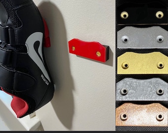 May include: A red plastic shoe hook with two gold screws is mounted on a white wall. A black cycling shoe with a white logo and the word "PELOTON" is hanging on the hook. The image also shows a variety of other colors for the shoe hook, including black, silver, gold, and brown.