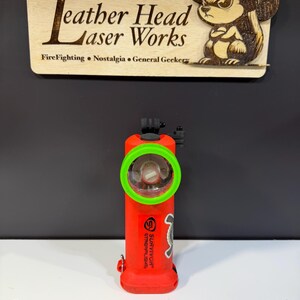 May include: A red flashlight with a green ring around the light. The flashlight has the word "SURVIVOR" and "STREAMLIGHT" printed on it. A wooden sign above the flashlight reads "Leather Head Laser Works" with a squirrel graphic.