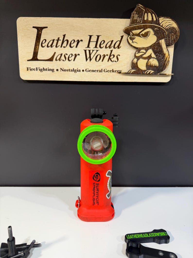 May include: A red flashlight with a green ring around the light. The flashlight has the word "SURVIVOR" and "STREAMLIGHT" printed on it. A wooden sign above the flashlight reads "Leather Head Laser Works" with a squirrel graphic.