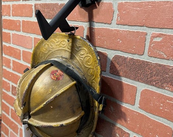 Op de afbeelding: Een zwarte metalen brandweerhelmhouder gemonteerd op een bakstenen muur. De houder is gevormd als een brandkraan en heeft een haak om een helm op te hangen. Een vintage gouden brandweerhelm hangt aan de haak.