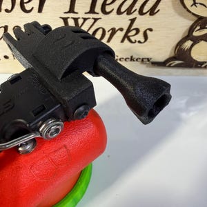 May include: A black 3D-printed device with a handle and a multi-pronged end, attached to a red and green object. The device is positioned in front of a wooden sign with the text "ber Head Works" and a cartoon bear.