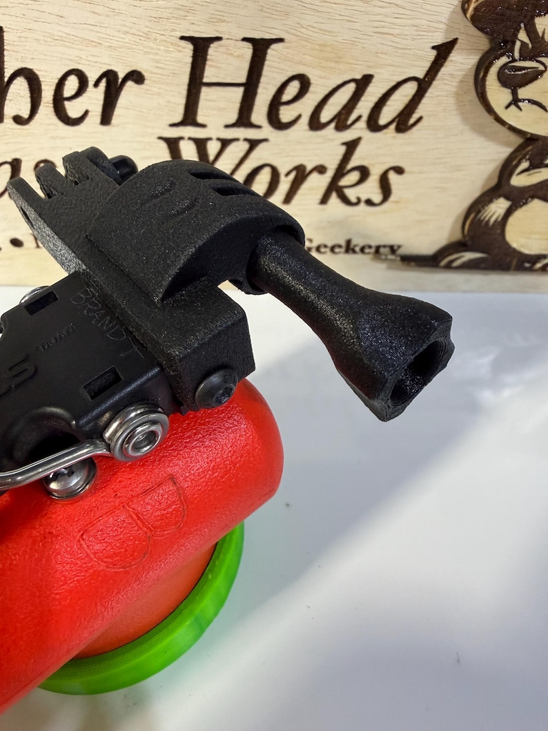 May include: A black 3D-printed device with a handle and a multi-pronged end, attached to a red and green object. The device is positioned in front of a wooden sign with the text "ber Head Works" and a cartoon bear.