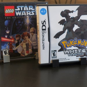 May include: Two Nintendo DS game cartridges, one for Lego Star Wars: The Original Trilogy and the other for Pokemon White Version. Both cartridges are displayed on a black surface with a black stand.