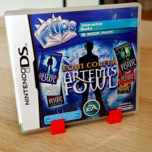 Könnte beinhalten: Eine Nintendo DS-Spielebox mit blauem und weißem Hintergrund. Das Spiel ist "Artemis Fowl" von Eoin Colfer. Die Box ist Teil eines 6-teiligen Pakets mit interaktiven Büchern. Die Box befindet sich auf einer Holzoberfläche.