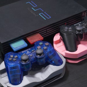 Könnte beinhalten: Eine schwarze PlayStation 2 Konsole mit zwei Controllern. Ein Controller ist schwarz und der andere ist blau. Beide Controller befinden sich in Ständern.