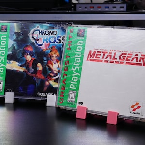 Puede incluir: Dos cajas de juegos de PlayStation con soportes de exhibición de plástico. Las cajas de juegos son verdes y blancas con el texto "Greatest Hits" y "PlayStation" en las espinas. Los títulos de los juegos son "Chrono Cross" y "Metal Gear Solid".