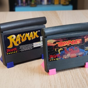 Könnte beinhalten: Zwei schwarze Atari Jaguar-Spielkassetten, eine für Rayman und eine für Tempest 2000. Die Rayman-Kassette hat ein gelbes und oranges Etikett mit dem Text "Rayman" und "Jaguar". Die Tempest 2000-Kassette hat ein rotes und oranges Etikett mit dem Text "Tempest 2000" und "Jaguar".