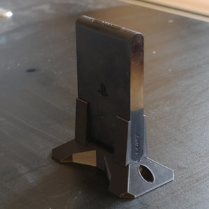 May include: A black plastic stand for a video game console. The stand has a PlayStation logo on the front and the word "TOPS" printed on the side.