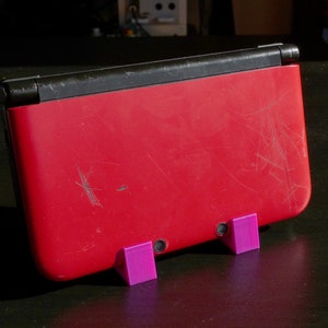 May include: A red Nintendo 3DS handheld gaming console with a black top and a scratched surface. The console is resting on two purple plastic stands.