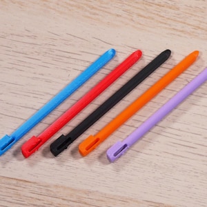 Puede incluir: Seis bolígrafos táctiles de diferentes colores, incluyendo azul, rojo, negro, naranja y morado, están dispuestos sobre una superficie de madera marrón claro.