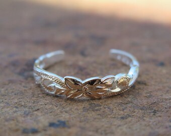 May include: A silver toe ring with a floral design. The ring is made of two bands, one with a smooth finish and the other with a textured finish. The floral design is in the center of the ring and is made of a different metal, possibly gold.