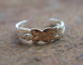May include: A silver and gold-toned adjustable toe ring with a floral design. The ring features a detailed flower in the center and ornate patterns along the band. The ring is designed to be worn on the toe.