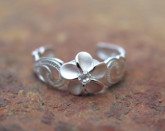 May include: A silver-colored toe ring featuring a flower design with a small, clear gemstone at the center. The ring has an open design and is decorated with swirling patterns. The ring is set against a brown, textured background.