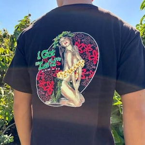 May include: Black t-shirt with a graphic on the back. The graphic shows a woman with flowers and a floral headpiece, set within a circular design with red and white floral accents. Text reads "I Got Lei'd" in green.