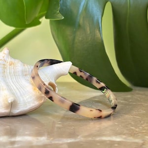 Può includere: Un braccialetto a cerchio con motivo tartarugato con macchie nere e beige. Il braccialetto è esposto vicino a una conchiglia bianca e foglie verdi. Il braccialetto è un accessorio elegante.