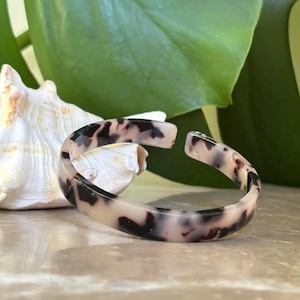 May include: Open-ended bangle bracelet with a tortoiseshell pattern in shades of cream and dark brown. The bracelet is curved and sits on a light-colored surface, with a seashell and green leaves in the background.