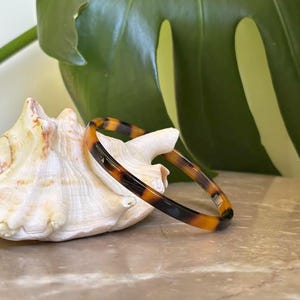 May include: A tortoiseshell-patterned bangle bracelet. The bracelet is a mix of brown, black, and amber colors. The bracelet is resting on a white seashell, with a large green leaf in the background.