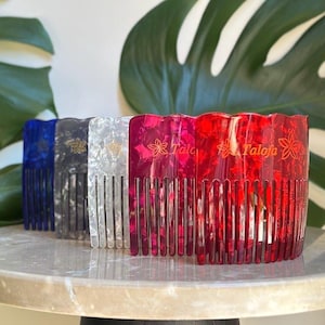Puede incluir: Una fila de cinco peines para el cabello coloridos en azul, negro, blanco, rosa y rojo. Cada peine tiene un borde superior festoneado y dientes verticales. El peine rojo tiene la palabra "Taloja" y un diseño floral.