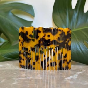 Puede incluir: Un peine con estampado de carey con un borde superior ondulado. El peine tiene forma rectangular con dientes muy juntos. El color es una mezcla de ámbar y marrón oscuro, con un acabado brillante. El peine es para el cuidado del cabello.