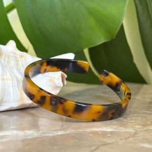 May include: A tortoiseshell-patterned cuff bracelet. The open-ended bangle features a warm palette of amber and dark brown hues. The bracelet is set against a backdrop of a seashell and a large green leaf.